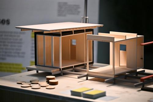 Architekturmodell aus hellem Holz auf einem Tisch: Ein moderner, modularer Baukörper mit Flachdach, großen Fensterflächen und offenem Eingangsbereich ist detailreich dargestellt; im Vordergrund liegen kleine Holzscheiben und Notizzettel.